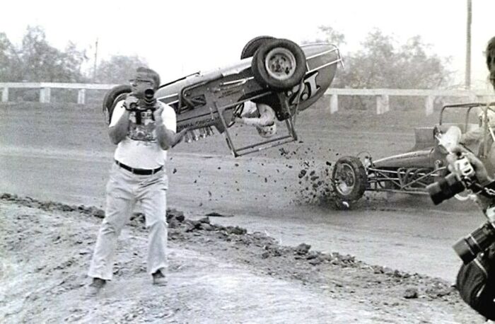 Black and white action photo capturing a perfectly timed crash at a dirt race with photographers in the frame.