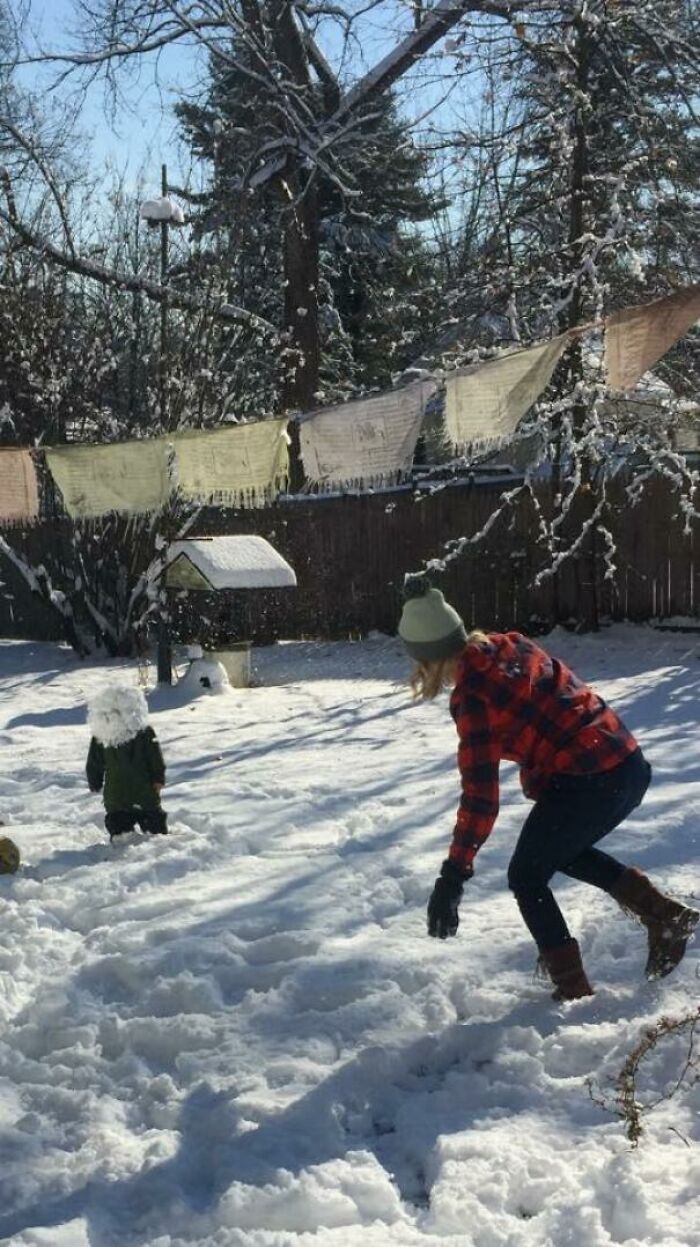 Child playing in snow with an adult wearing a green hat and red plaid jacket in a snowy backyard scene, perfect timed photo.