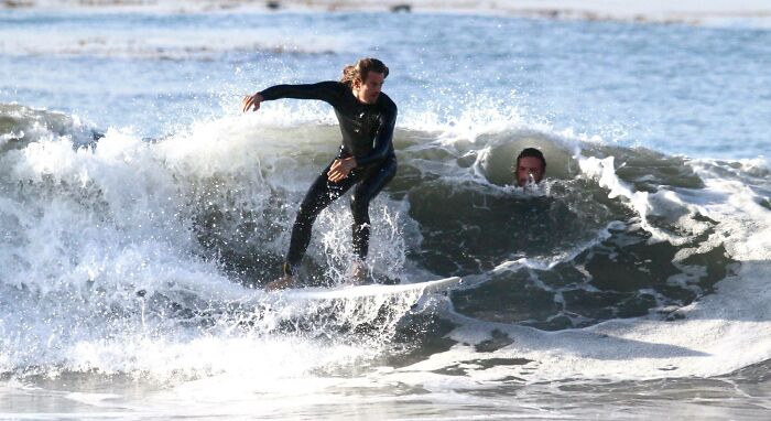Surfer riding a wave perfectly timed with another person’s head appearing inside the curling water splash.