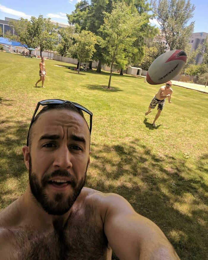 Man taking a perfectly timed photo as a rugby ball appears to hit his head in a sunny park setting.