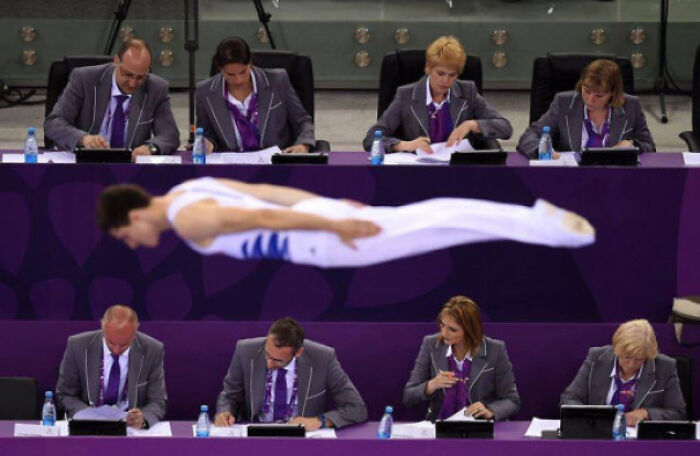 Gymnast captured in a perfectly timed photo mid-air during a routine, with judges focused on scoring and notes.