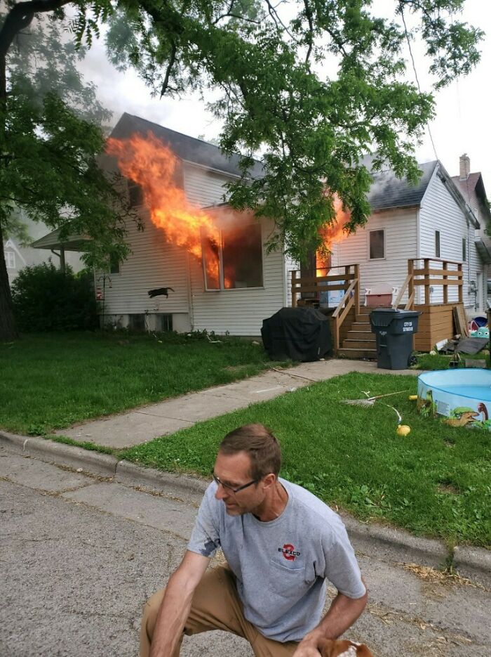 Man crouching on the street with a house on fire in the background captured in a perfectly timed photo.