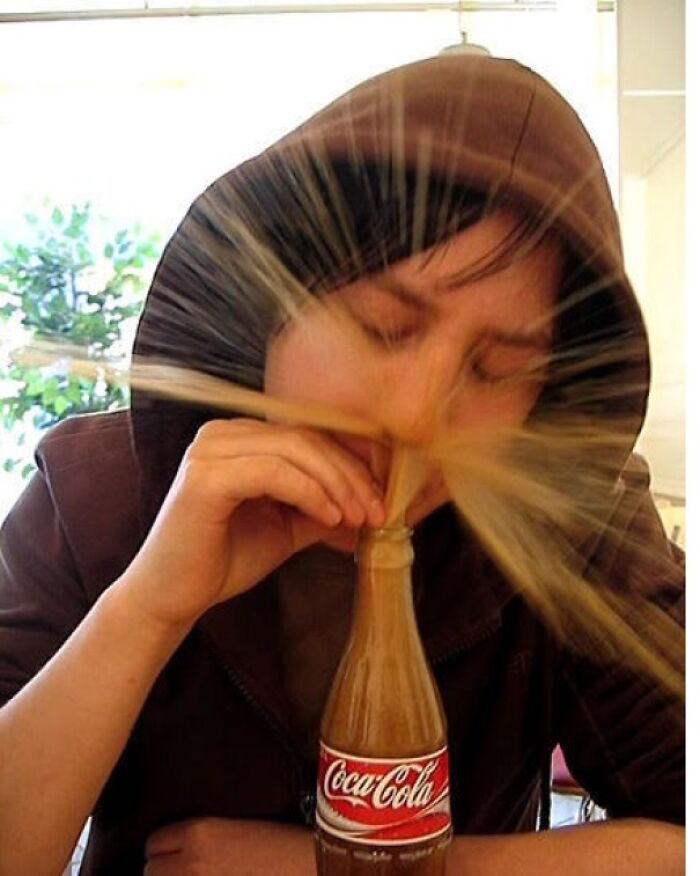 Person in brown hoodie accidentally sprays soda from bottle, captured in a perfectly timed photo showing a burst of liquid.
