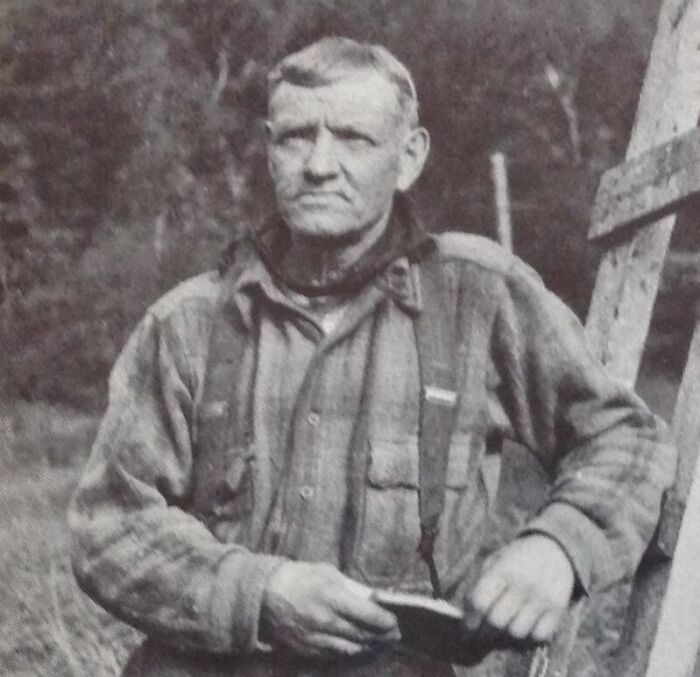 Black and white photo of an American folk hero in rugged clothing holding a tool outdoors near a wooden ladder.
