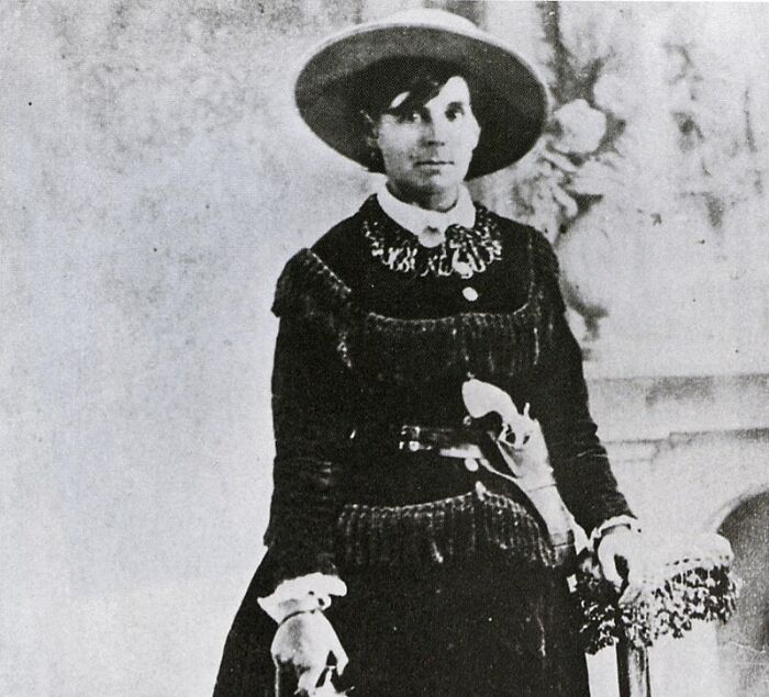 Black and white photo of an American folk hero woman in Western attire and wide-brimmed hat holding a rifle.