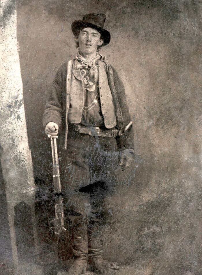 Faded sepia image of a 19th-century American folk hero standing with rifle in rustic frontier clothing and hat.