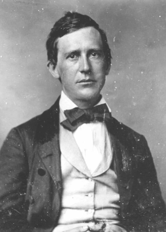 Black and white portrait of a 19th-century American folk hero wearing a suit and bow tie, looking directly at the camera