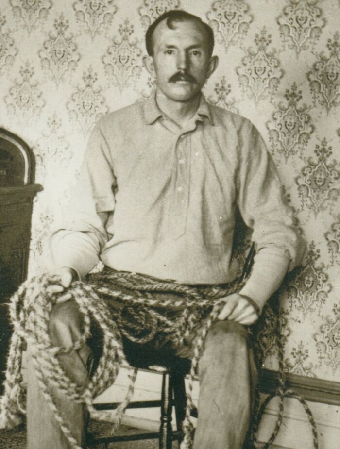 Vintage photo of an American folk hero sitting on a chair wrapped with rope, showcasing early 20th-century attire and setting.