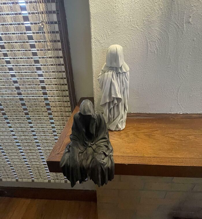 Two ghost-like statues with hooded cloaks, one black and one white, placed on a wooden shelf near a window blind.