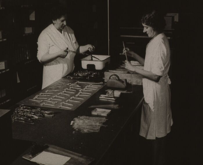 Two women in vintage medical uniforms preparing instruments in an old-school medical setting with sterilization tools.