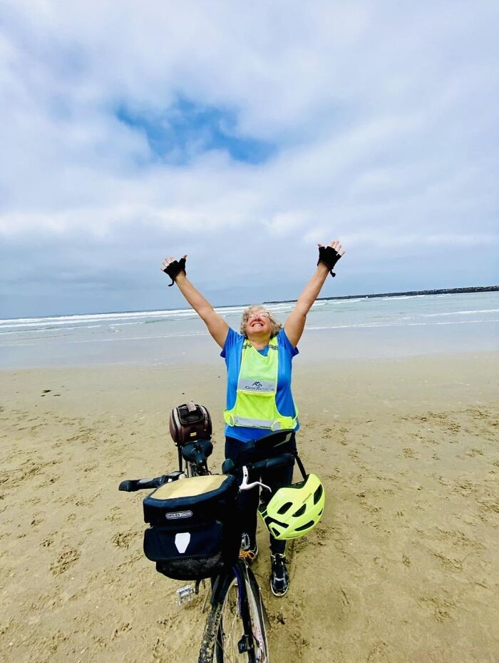 Person celebrating on beach with bicycle and helmet, enjoying a wholesome moment by the ocean under a cloudy sky.