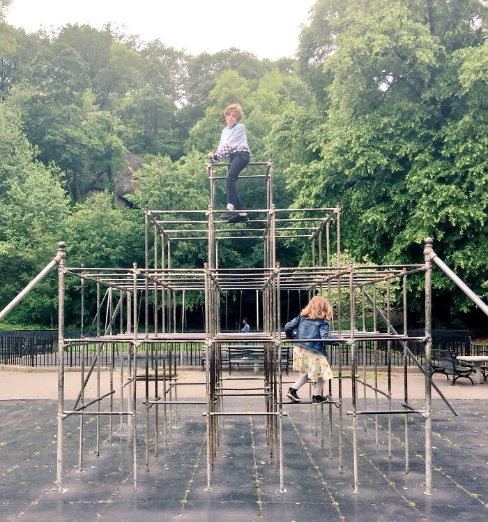 Children climbing on a vintage metal playground structure in a park surrounded by lush green trees.