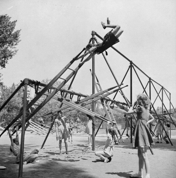 Historic playground with children climbing and swinging on risky metal structures in a black and white photo.