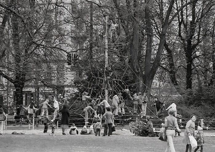 Historic photo of playground with many children climbing risky rope structures surrounded by trees and adults nearby.