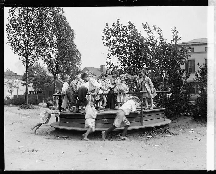 Historic playground photo showing children playing on a wooden merry-go-round in an early 20th century park.