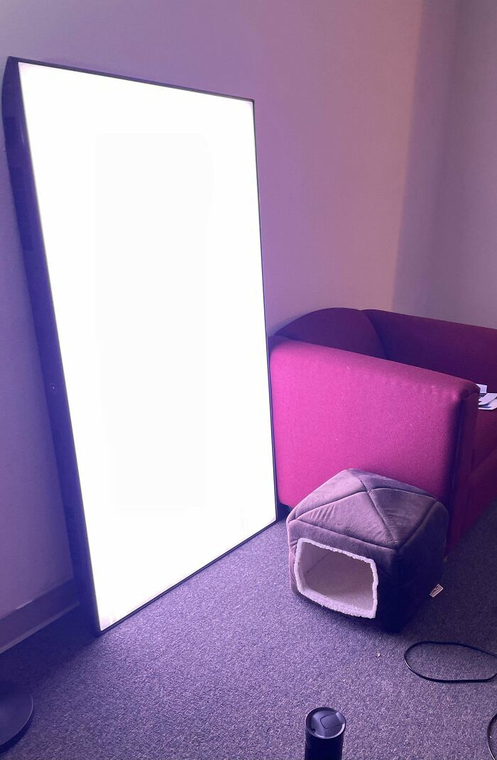 Large illuminated panel leaning against a wall beside a red couch and a small plush pet cube in a carpeted room.