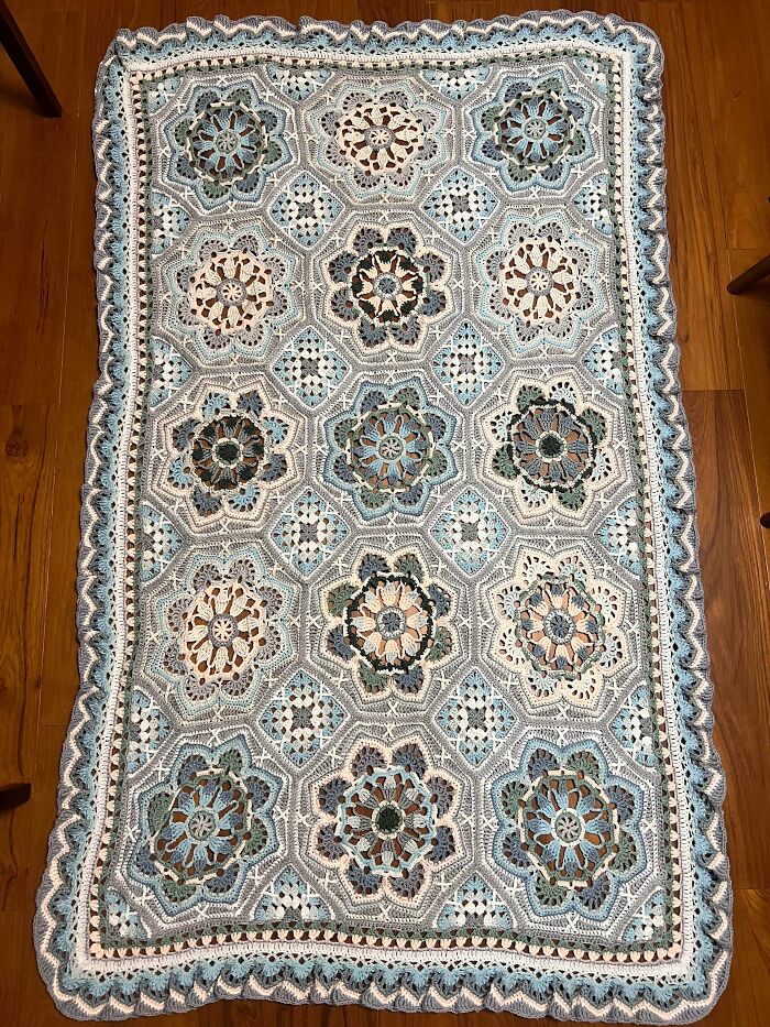 Intricate crocheted blanket with floral motifs and geometric patterns in soft blue, beige, and white colors on wood floor.