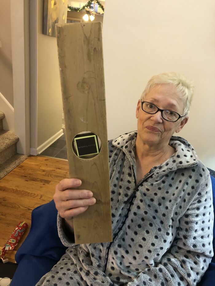 Elderly woman in polka dot robe holding a plank of wood with a hole, showing a humorous parent gift idea.