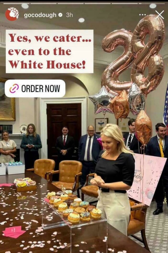 Woman smiling at decorated donuts on table during chaotic good celebration with balloons and people in a formal room.