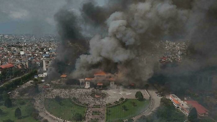 Aerial view of a chaotic scene with thick smoke rising over a large building surrounded by a crowd in a city park.