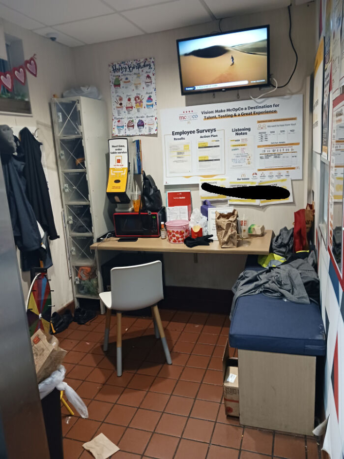 Cluttered and cramped break room with tiled floor, small table, single chair, microwave, and wall-mounted TV, resembling a prison setting.
