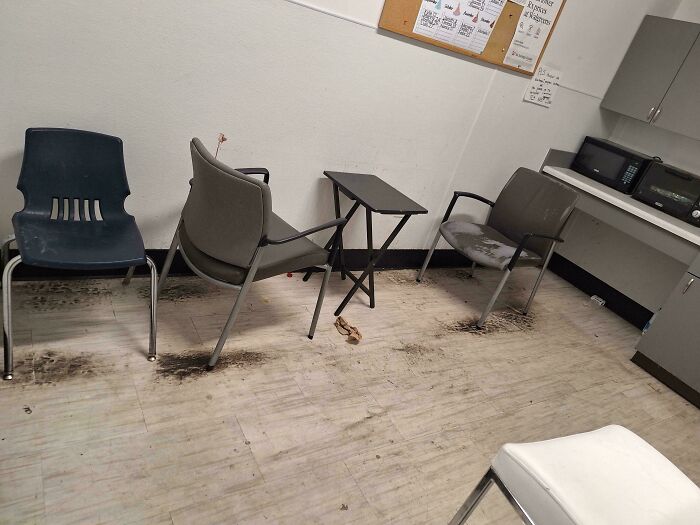Dirty break room with stained floor and worn chairs resembling a prison setting with minimal furniture and appliances.