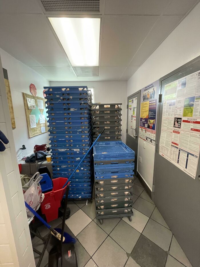 Cluttered break room with stacks of trays, cleaning supplies, and posted notices, resembling a prison-like atmosphere.