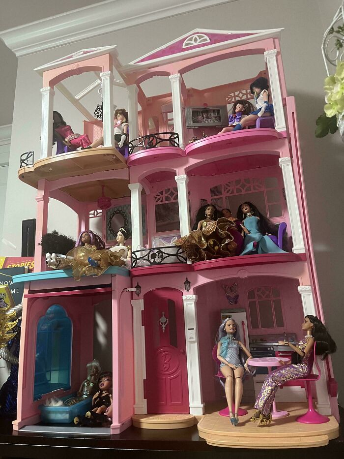 Pink dollhouse with multiple levels and dolls dressed in elegant outfits in various rooms and balconies.