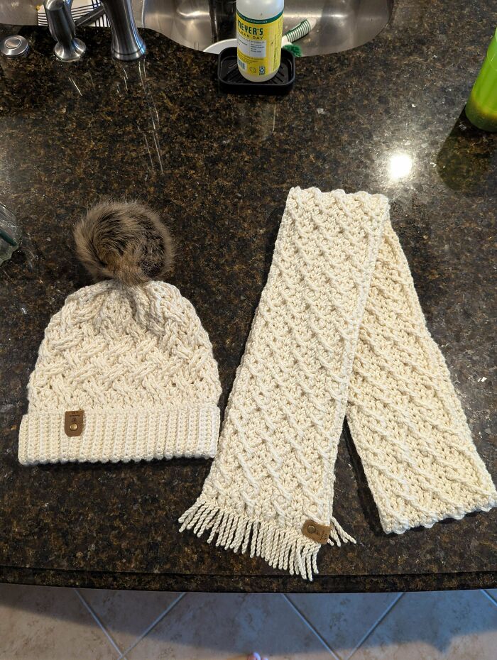 Crocheted cream-colored hat with fur pom-pom and matching textured scarf laid out on dark kitchen countertop.