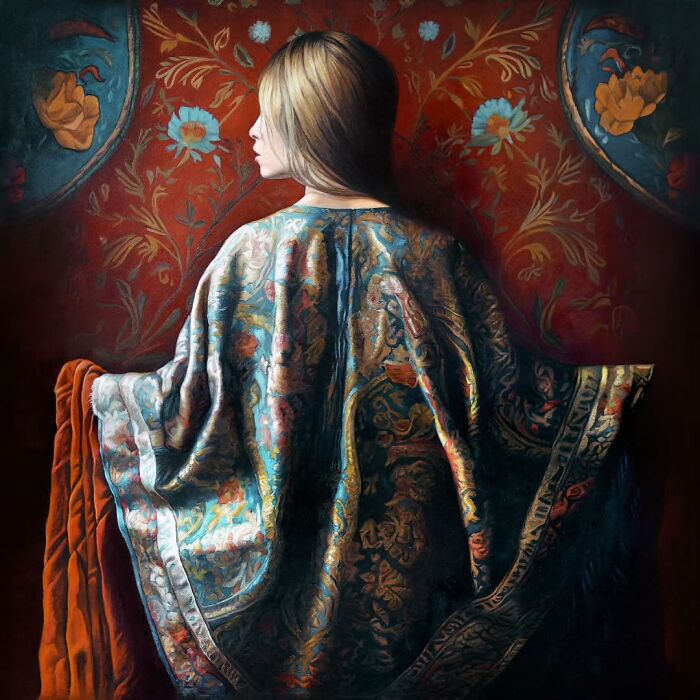 Woman draped in colorful fabric stands against an ornate red patterned background, celebrating women’s strength and beauty.