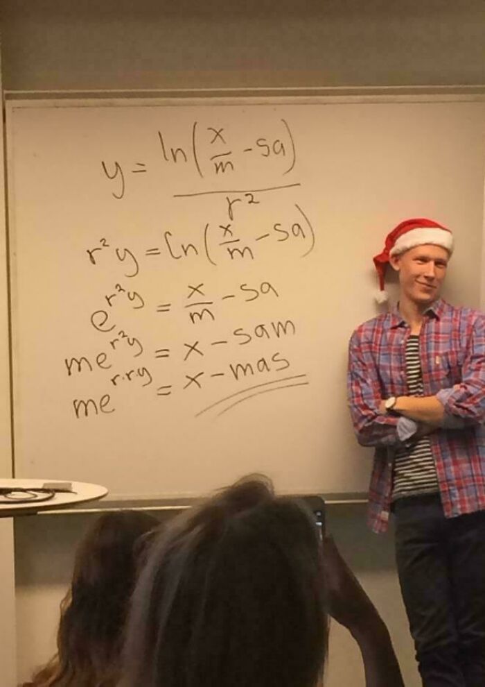 Teacher wearing a Santa hat standing beside a whiteboard with a funny math equation spelling out "x-mas" for humor.