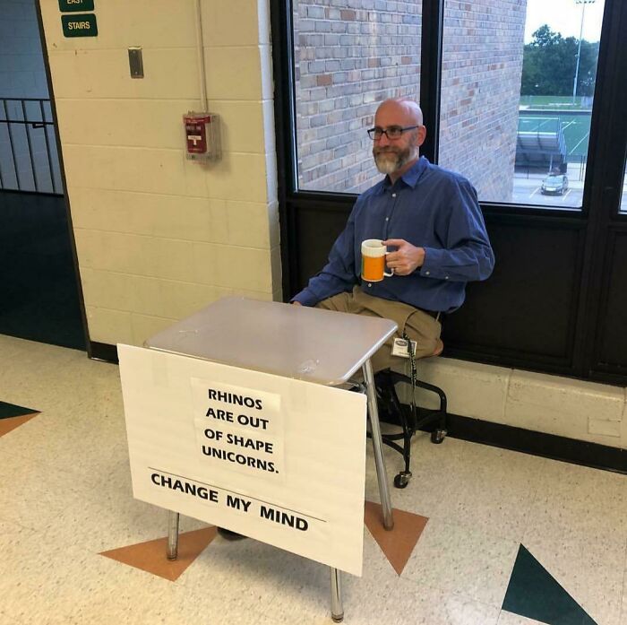 Male teacher with a beard sitting at a desk holding a coffee mug, displaying a funny sign about rhinos and unicorns.