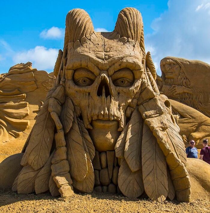 Intricate sand sculpture of a skull with feathers, showcasing Danish artistic creativity under a clear blue sky.