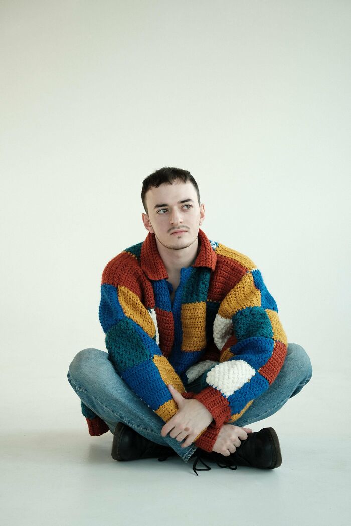 Young man wearing a colorful crocheted sweater sitting cross-legged on the floor, showcasing crocheting craftsmanship.