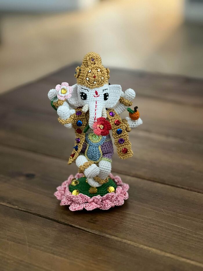 Crocheted figurine of an elephant deity adorned with colorful decorations standing on a pink crocheted base.