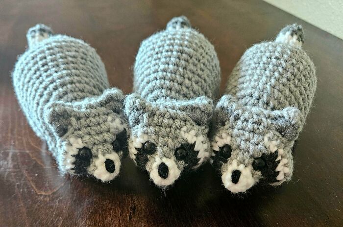 Three crocheted raccoons in gray yarn displayed on a wooden surface, showcasing detailed handcrafted crochet art.