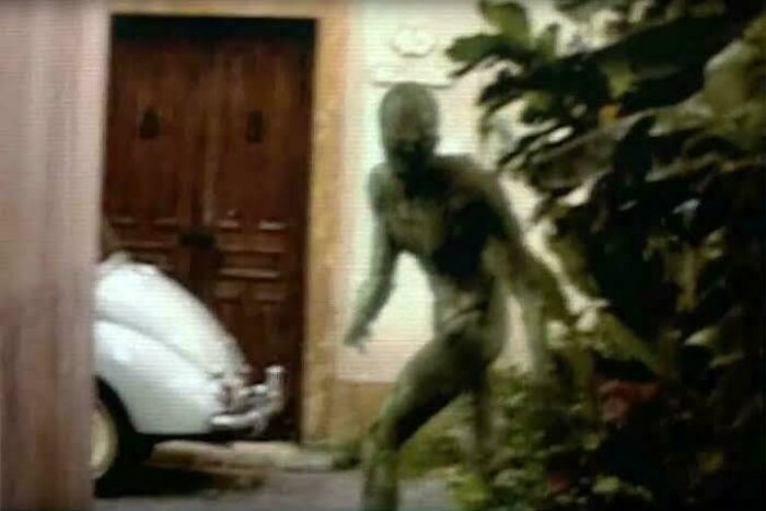 A blurry, green, humanoid figure emerging from behind lush foliage near a vintage white car and wooden door, a creepy image.