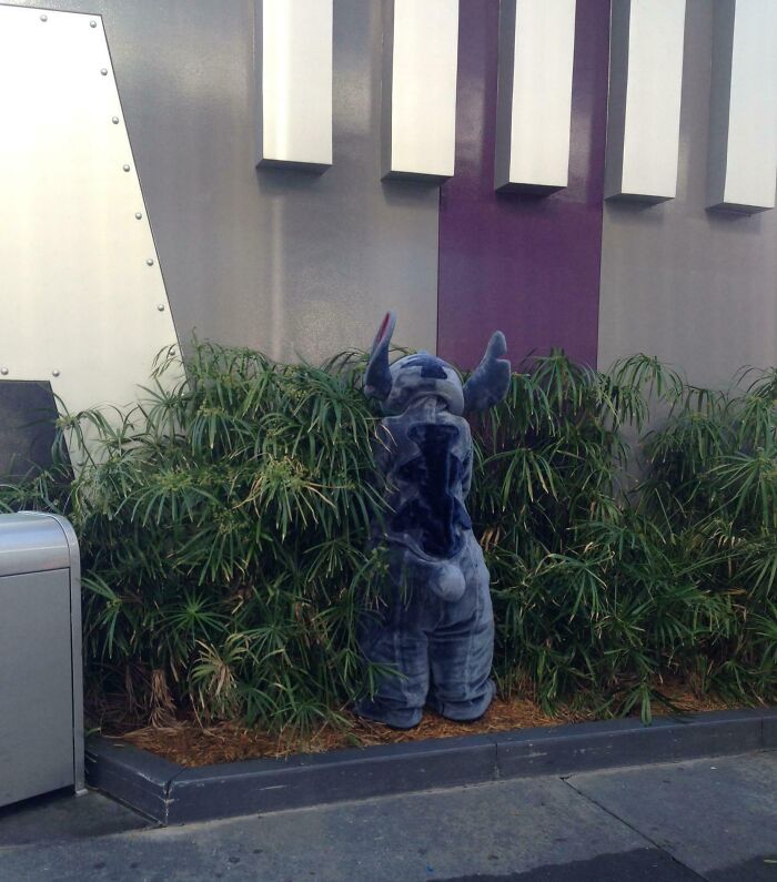 Person in a Stitch costume standing with their back to the camera near greenery at an amusement park.