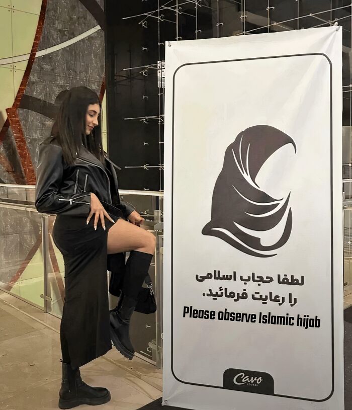 Woman wearing a black leather jacket and boots posing defiantly next to a sign about observing Islamic hijab rules.