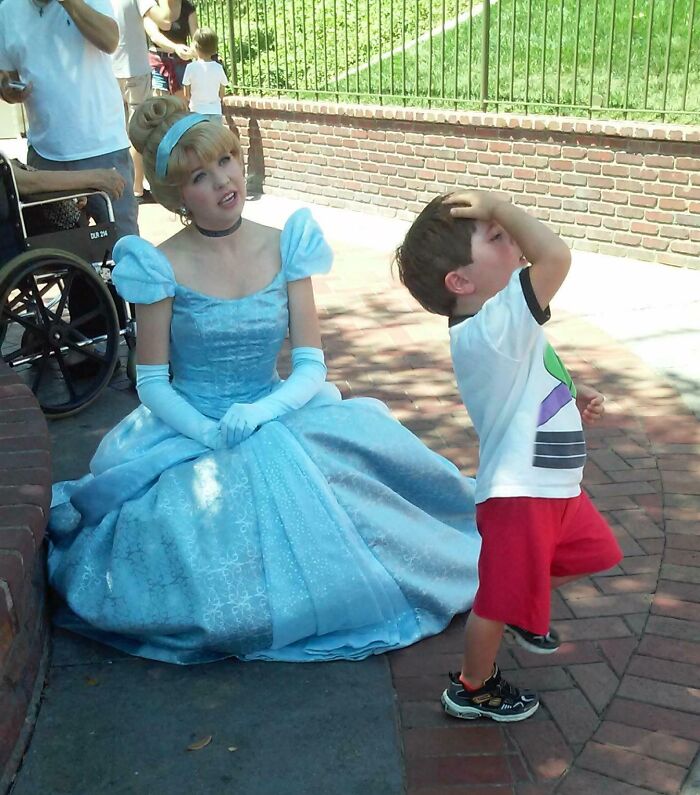 Woman dressed as a princess sitting on the ground while a young boy stands nearby at an amusement park.