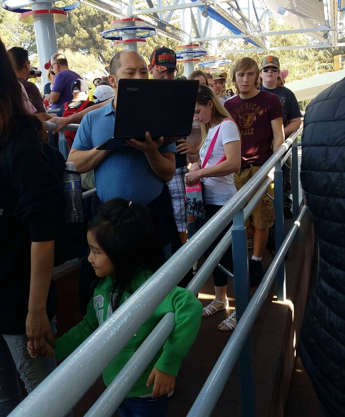 Man using laptop in a crowded amusement park queue with families waiting to enjoy the rides on a sunny day.
