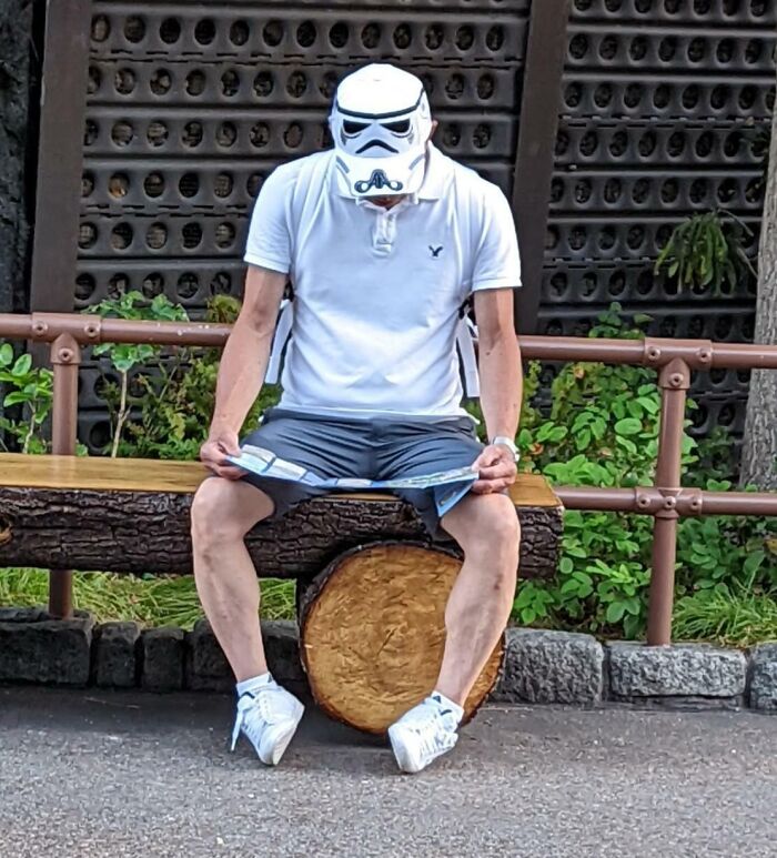 Person wearing a Stormtrooper hat sitting on a log bench, holding a map at an amusement park visit.