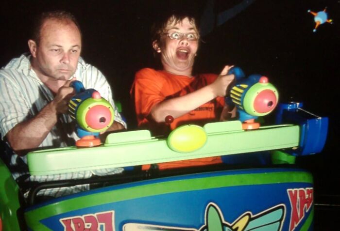 Man and excited boy riding and aiming laser guns on an amusement park ride during a nighttime game.