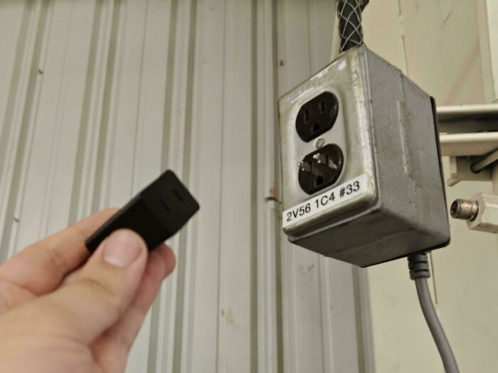Hand holding a small black adapter plug near a metal electrical outlet box in an industrial setting, moderately interesting.