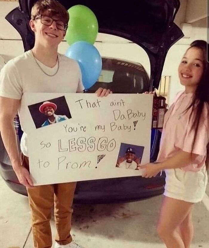 Young couple holding a cringe-worthy but wholesome promposal sign with playful text and balloons in the background.