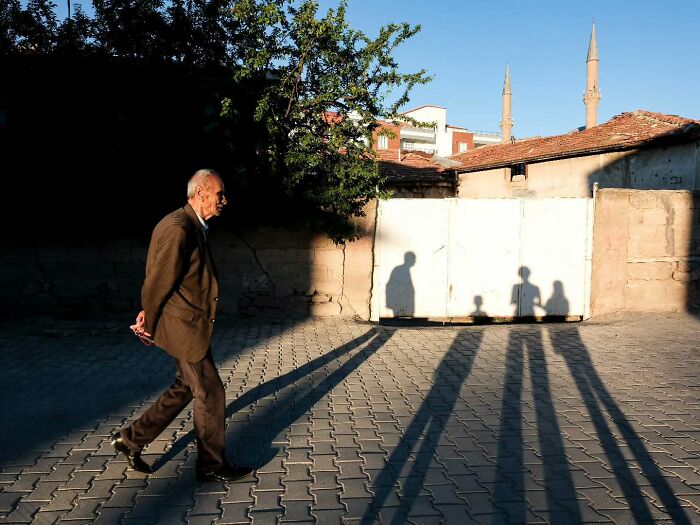Elderly man walking on street with long shadows cast on pavement in a perfectly timed street photo by Melahat Ünel.