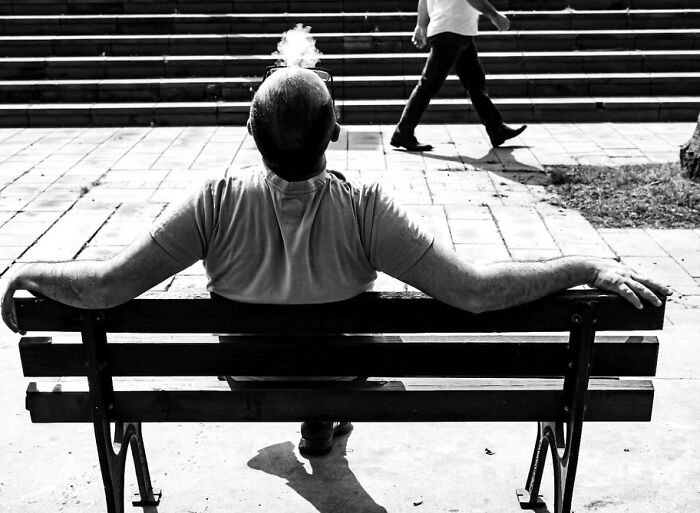 Man sitting on a park bench smoking a cigarette as a pedestrian walks by, captured in a perfectly timed street photo.