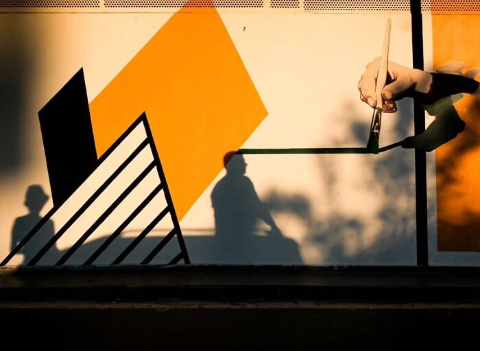Shadow of a person with a hat blends into a street mural with geometric shapes in perfectly timed street photography.