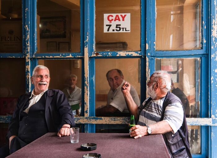 Two elderly men sharing a laugh and drink at a cafe, captured in a perfectly timed street photo by Melahat Ünel.