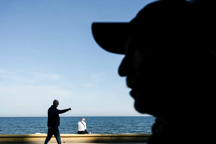 Silhouette of a man wearing a cap with two people near the sea in a perfectly timed street photo by Melahat Ünel.